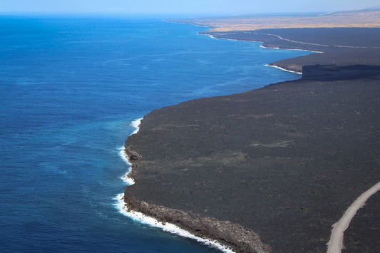 A dramatic aerial view of a black lava flow on the Island of Hawaii, with the ocean to the left and a road cutting through the landscape.