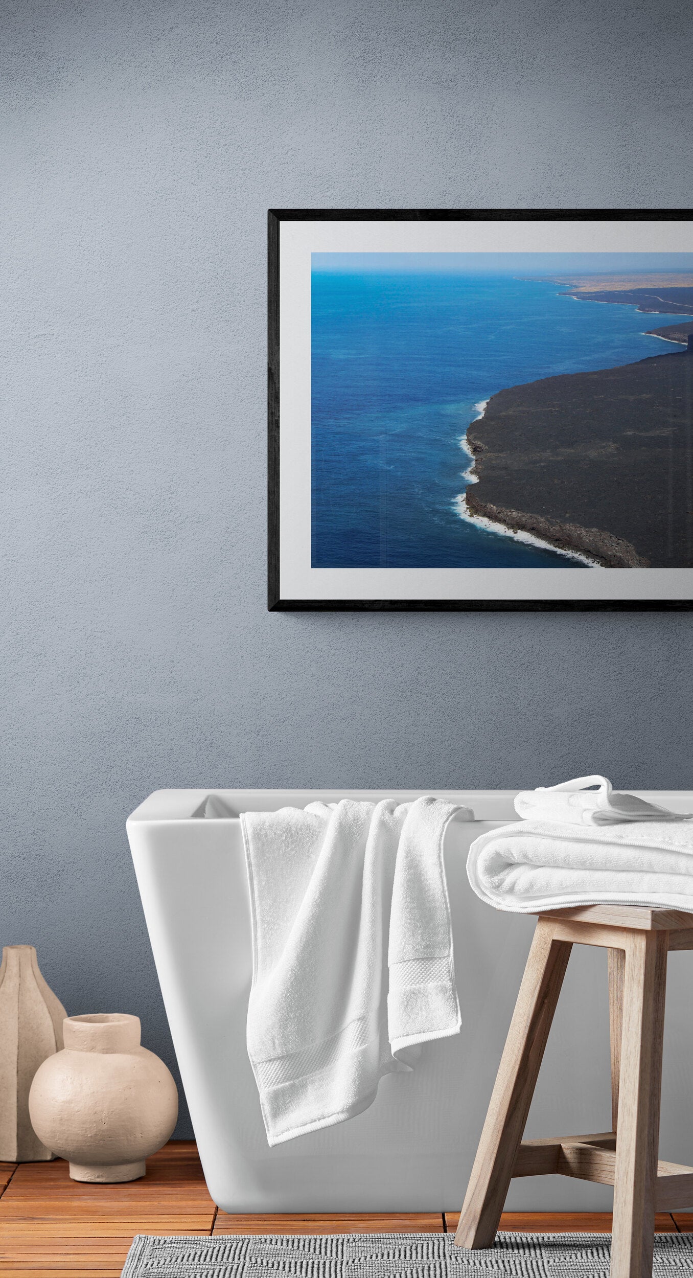 Framed picture of a coastal landscape above a white bathtub with towels.