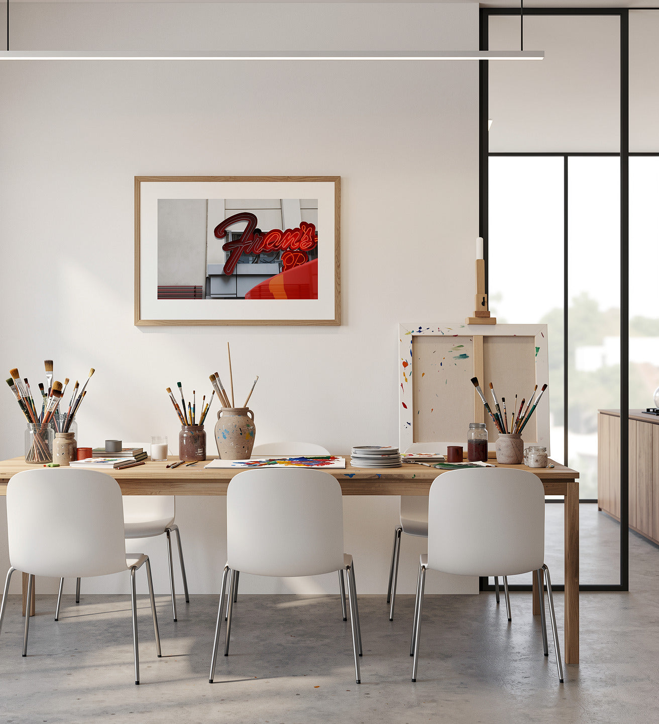 A framed art print featuring a red and black abstract design hangs on a white wall in a modern, sunlit artist's studio. The room is styled with a wooden table, white chairs, paintbrushes in jars, and a small easel with colorful paint splatters.