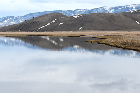 A color photograph of Jackson Watering Hole, featuring a small lake with a reflection of mountains in the water, surrounded by brown grasses and a backdrop of snow-capped mountains under a cloudy sky.