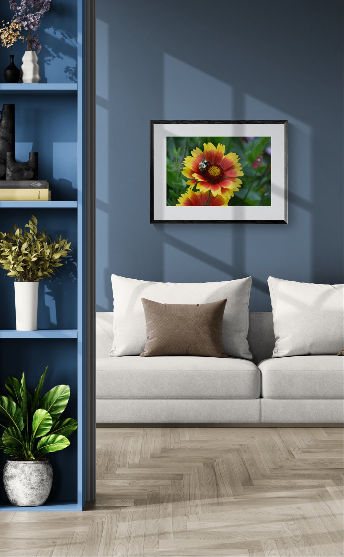 A framed floral art print featuring yellow and orange flowers with green leaves, displayed on a dark blue wall above a light gray sofa in a modern living room setting.