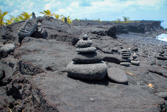 Stacked rocks on a black sand beach with lava-scarred earth and resilient trees in the background, including a large rock with a carved face mask.
