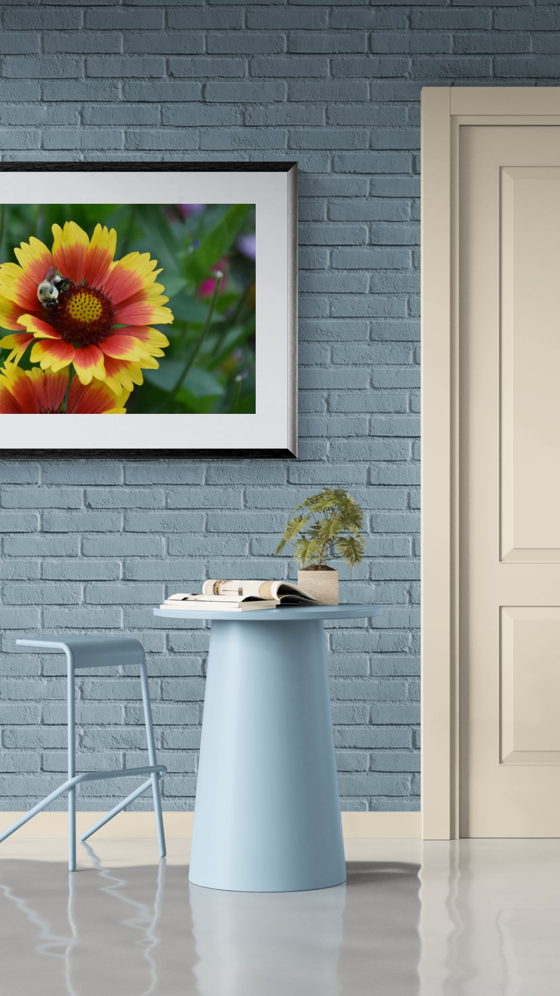 A framed wall art print of a yellow and red flower with a bee, displayed on a blue brick wall above a light blue table in a modern interior setting.