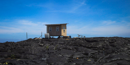 A panoramic photograph featuring a small wooden shack on a rugged, lava-covered landscape with a clear blue sky in the background. A water tank and a patio lounge chair are visible in front of the shack.