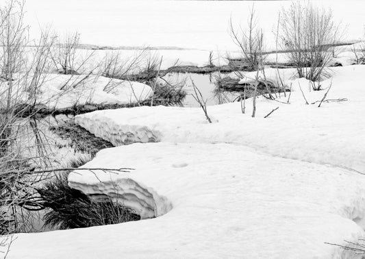 A black and white photograph of a serene winter scene with snow on the ground and a partially frozen creek, with footprints or marks on the snow surface.