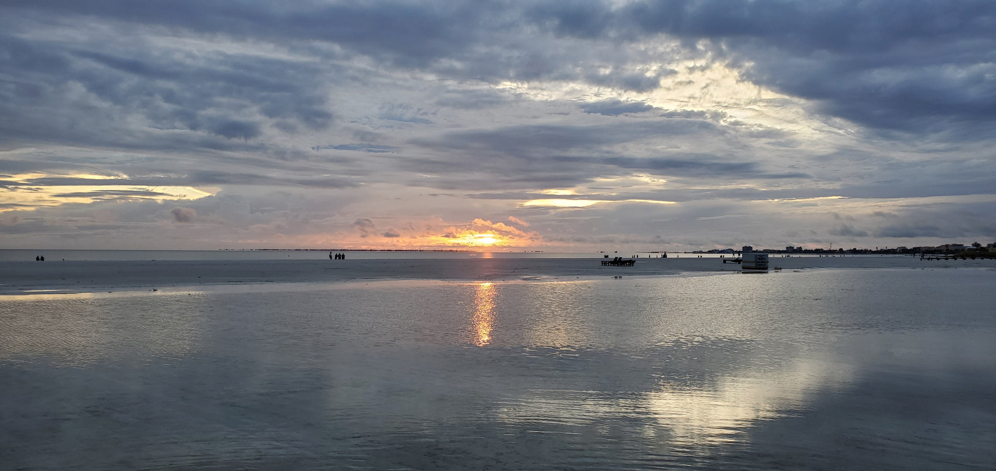 A photograph of a sunset at Ft. Myers Beach, featuring the sun low in the sky with a reflective body of water and a blue, cloudy sky. Sunset at Ft. Myers Beach, Original Photograph by Kim A. Bailey