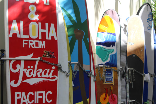 A photograph featuring multiple surfboards leaning against a wall in Honolulu, Hawaii, with a red sign that reads 'Aloha from Ökai Pacific' in the foreground. Surfboards in Honolulu, Hawaii, Original Photograph by Kim A. Bailey
