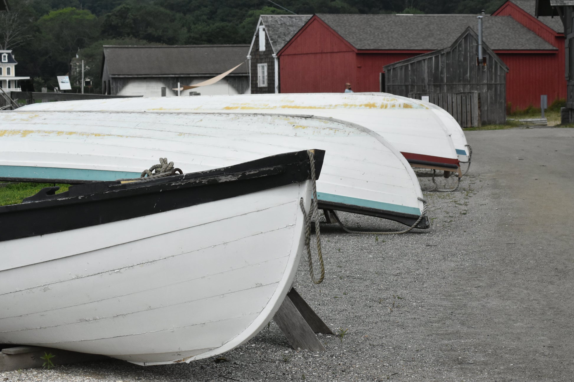 Row of white boats on a gravel surface with red buildings in the background. Wooden Boats Original Photograph by Kim A. Bailey